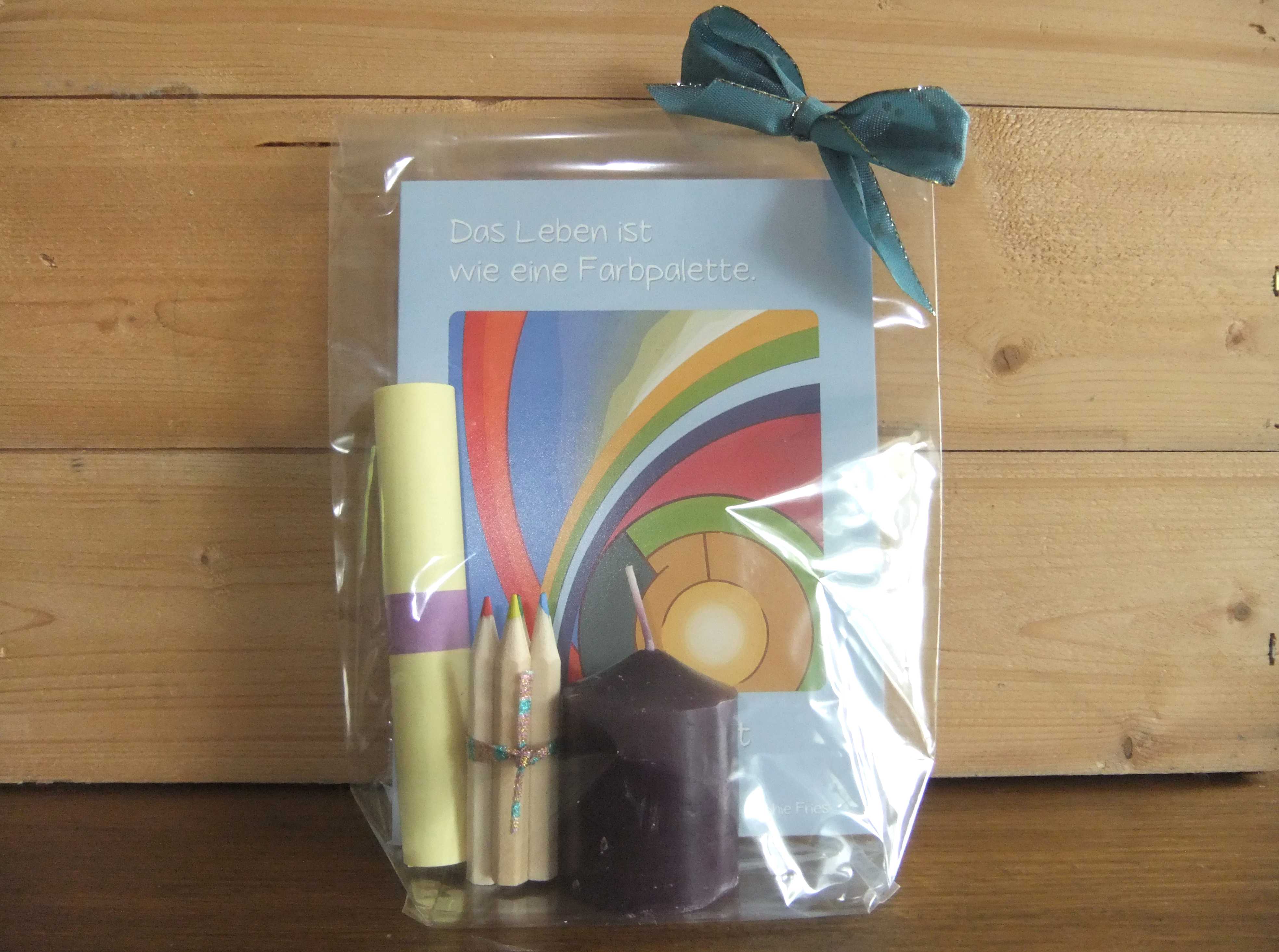 Geschenkig verpackte Faltkarte mit farbenfreudigem Regenbogen vor hellblauem Rahmen, dazu eine Kerze, Mini-Buntstifte und ein Text.